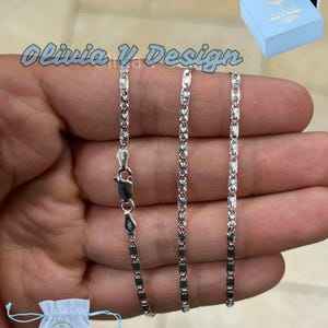 May include: Three silver chains with a diamond-shaped pattern. The chains are on a hand and are all the same length. The chains are in a white bag with the text "Olivia V Design" on it.