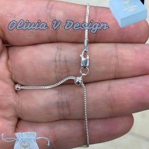 May include: A silver chain bracelet with a sliding clasp and two small silver beads. The bracelet is on a hand and the text "Olivia V Design" is visible in the background.