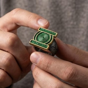 May include: A ring featuring a prominent green circular emblem with a central spiral design, framed by a gold-toned rectangular border. The band of the ring is dark gray with a textured, cracked surface finish, contrasting with the bright green inlay.