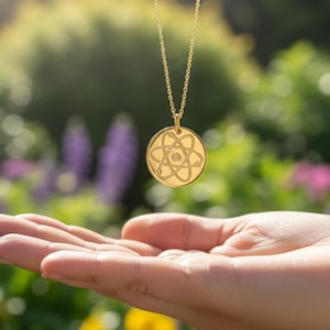 May include: A gold-colored pendant necklace with a circular charm featuring an atom design. The necklace hangs above an open hand, set against a blurred background of green foliage and colorful flowers.