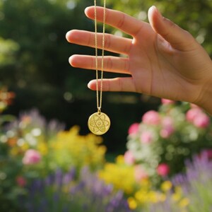 May include: A gold-colored pendant necklace with a circular charm featuring a geometric design. The necklace is held up against a blurred background of colorful flowers and greenery.