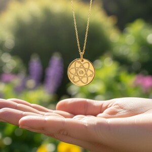 May include: A gold-colored pendant necklace with a circular charm featuring an atom-like design. The necklace hangs above an open hand, set against a blurred background of green foliage and colorful flowers.