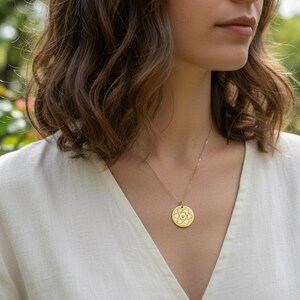 May include: A gold pendant necklace with a circular charm featuring an intricate floral design. The necklace hangs on a delicate chain against a cream-colored top. The background shows a garden with pink and yellow roses.