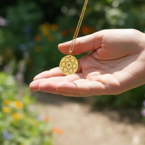 May include: A gold-colored necklace with a pendant featuring an atom design. The necklace is suspended from a delicate chain and held in an open hand against a blurred background of greenery and flowers.