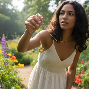 May include: A woman in a white dress holds a gold pendant necklace in a garden setting. The necklace features a circular pendant with an intricate design. The background includes colorful flowers and greenery, creating a natural and serene atmosphere.