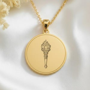 Op de afbeelding: Een goudkleurige, ronde hanger aan een ketting met een centrale gravure van een brandende fakkel. De hanger hangt aan een fijne goudkleurige ketting en heeft een gepolijste metalen afwerking en een strak, cirkelvormig ontwerp.