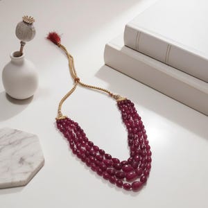Könnte beinhalten: Eine mehrsträngige Halskette mit tiefroten Perlen. Die Halskette hat ein goldfarbenes, verstellbares Band und eine rote Quaste. Der Schmuck wird auf einer weißen Oberfläche präsentiert, zusammen mit einer kleinen weißen Vase mit einer getrockneten Blume und einem Stapel Bücher.