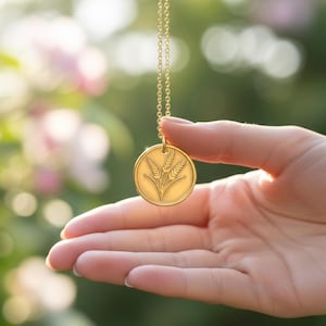 Puede incluir: Un collar con colgante dorado con un dije circular que representa trigo. El collar se sostiene en una mano sobre un fondo borroso de follaje verde y rosa, lo que sugiere un entorno al aire libre.