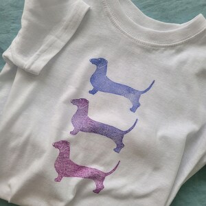 Puede incluir: Camiseta blanca con tres siluetas de perro salchicha en tonos morados y azules. Los perros están estampados en la parte delantera de la camiseta. La camiseta tiene mangas cortas y cuello redondo.