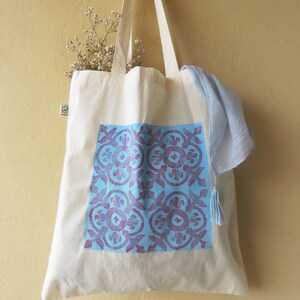 May include: A white canvas tote bag with blue and purple floral pattern printed on the front. The bag has a light blue fabric strap and a tassel.