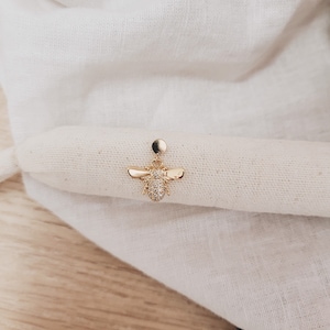 Può includere: Un orecchino a forma di ape dorata con accenti di cristallo trasparente. L'orecchino è su uno sfondo di tessuto bianco.