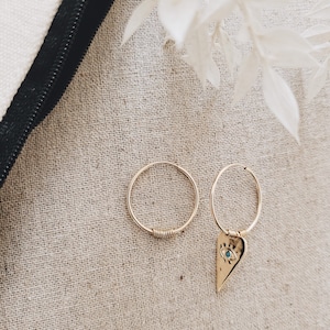 May include: Two gold hoop earrings, one with a small heart charm featuring an evil eye design. The earrings are displayed on a beige fabric background.
