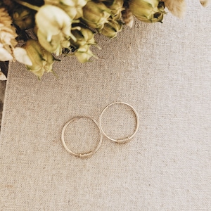 Puede incluir: Dos pendientes de aro dorados sobre un fondo de tela beige. Los aros son simples y delicados, con un diseño de alambre ligeramente retorcido.