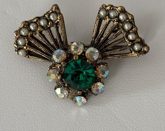 May include: A gold tone brooch with a green rhinestone center surrounded by smaller clear rhinestones. The brooch has two wings with pearl accents.