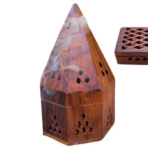 May include: A wooden incense burner with a pyramid shape and intricate carvings. The burner has a square base and a pointed top. Smoke is rising from the top of the burner.