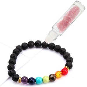 May include: A black lava rock bracelet with gold beads and seven chakra stones. A clear glass roller bottle filled with pink crystals is being used to add essential oils to the bracelet.