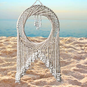 May include: A handcrafted, circular wall hanging made of natural seashells and beads. The design features a spiral pattern with dangling shell accents, creating a coastal, bohemian aesthetic. The artwork is displayed on a sandy beach with the ocean in the background.