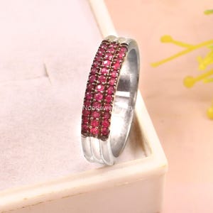 Puede incluir: Un anillo plateado con tres filas de pequeñas gemas rojas vibrantes. El anillo se presenta sobre un fondo rosa pálido, con un toque de vegetación verde. El diseño es sencillo, resaltando el rico color de las gemas.