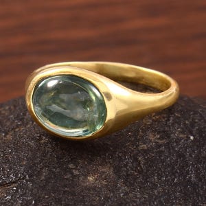 Pode incluir: Um anel dourado com uma grande pedra preciosa ovalada verde claro. O anel está sobre uma superfície escura e texturizada, com um fundo de madeira. A banda do anel é lisa e polida.