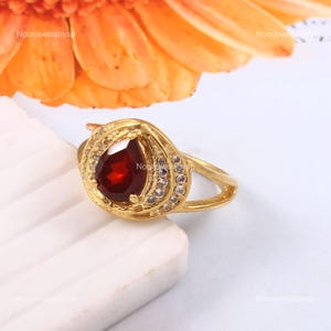 Op de afbeelding: Een gouden ring met een peervormige, dieprode edelsteen, omringd door een halo van kleine, heldere stenen. De ring heeft een delicaat, gedraaid ontwerp. De ring staat tegen een witte en blauwe achtergrond, met een oranje bloem op de achtergrond.