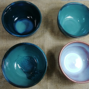 Op de afbeelding: Vier keramische kommen in verschillende tinten blauw en groen met een witte glazuur. De kommen zijn gerangschikt op een jute oppervlak.