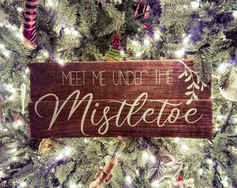 May include: A rustic wooden sign with the text "Meet me under the mistletoe" in white lettering. The sign is hanging on a Christmas tree decorated with lights and ornaments.