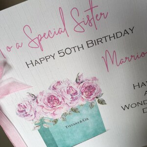 Puede incluir: Tarjeta de felicitaci&oacute;n con el texto "To a Special Sister HAPPY 50TH BIRTHDAY". La tarjeta presenta un ramo de rosas rosas y una ilustraci&oacute;n de una caja Tiffany & Co. Una cinta rosa est&aacute; atada a la tarjeta.