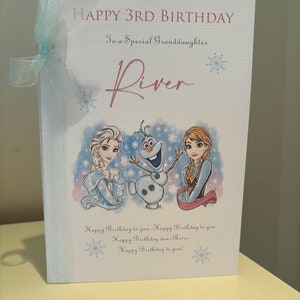 K&ouml;nnte beinhalten: Geburtstagskarte mit dem Text "HAPPY 3RD BIRTHDAY" und "To a Special Granddaughter River". Die Karte zeigt Illustrationen von Figuren aus dem Film Frozen und ist mit einem hellblauen Band gebunden.
