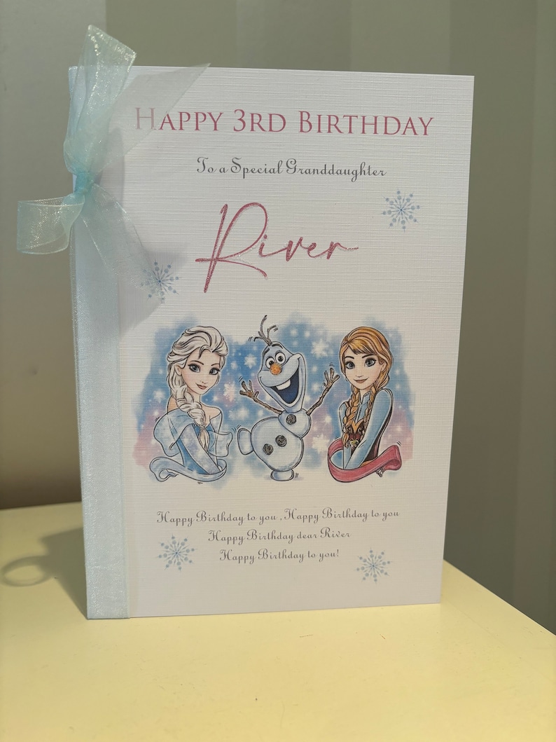 K&ouml;nnte beinhalten: Geburtstagskarte mit dem Text "HAPPY 3RD BIRTHDAY" und "To a Special Granddaughter River". Die Karte zeigt Illustrationen von Figuren aus dem Film Frozen und ist mit einem hellblauen Band gebunden.