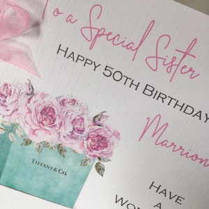 Puede incluir: Una tarjeta de felicitaci&oacute;n con el texto "To a Special Sister HAPPY 50TH BIRTHDAY" en escritura rosa. La tarjeta presenta un dise&ntilde;o floral con rosas rosas y una caja Tiffany & Co. Una cinta rosa est&aacute; atada en la parte superior izquierda.