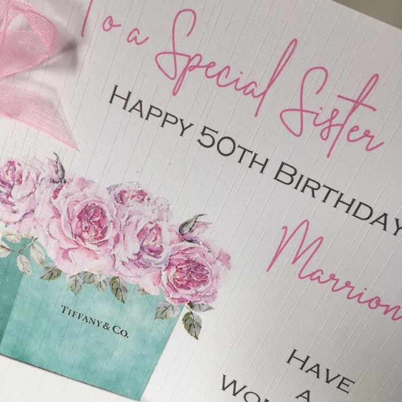 Puede incluir: Una tarjeta de felicitaci&oacute;n con el texto "To a Special Sister HAPPY 50TH BIRTHDAY" en escritura rosa. La tarjeta presenta un dise&ntilde;o floral con rosas rosas y una caja Tiffany & Co. Una cinta rosa est&aacute; atada en la parte superior izquierda.