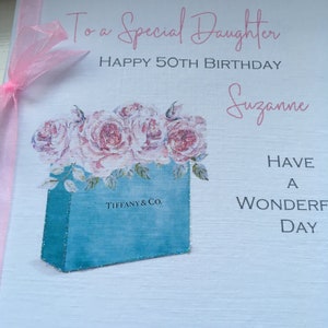 Puede incluir: Una tarjeta de felicitaci&oacute;n con el texto "To a Special Daughter, Happy 50th Birthday, Suzanne". Presenta una bolsa de regalo azul con el logo de Tiffany & Co., llena de rosas rosas. Una cinta rosa est&aacute; atada al lado izquierdo.