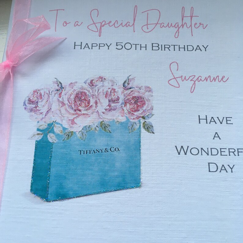 Puede incluir: Una tarjeta de felicitaci&oacute;n con el texto "To a Special Daughter, Happy 50th Birthday, Suzanne". Presenta una bolsa de regalo azul con el logo de Tiffany & Co., llena de rosas rosas. Una cinta rosa est&aacute; atada al lado izquierdo.