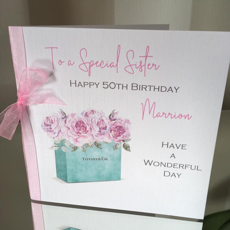 Puede incluir: Tarjeta de cumplea&ntilde;os con el texto "To a Special Sister, Happy 50th Birthday Marrion". La tarjeta presenta una ilustraci&oacute;n de acuarela de rosas rosas en una caja Tiffany & Co., con una cinta rosa.