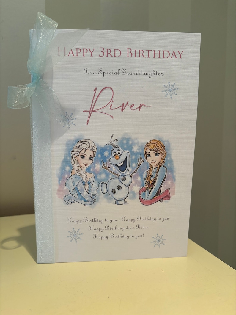K&ouml;nnte beinhalten: Geburtstagskarte mit hellblauem Band. Auf der Karte steht "HAPPY 3RD BIRTHDAY" und "To a Special Granddaughter River". Sie zeigt Illustrationen von Elsa, Olaf und Anna aus Frozen. Auf der Unterseite der Karte steht "Happy Birthday to you!"