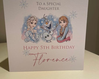 以下が含まれることがあります： 「特別な娘へ」と「5歳の誕生日おめでとう Florence」の文字が入った、薄いピンク色の誕生日カード。映画「アナと雪の女王」のエルサ、アナ、オラフのイラストが描かれており、雪の結晶のアクセントが施されています。