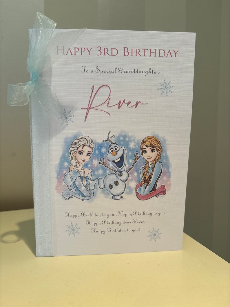 K&ouml;nnte beinhalten: Geburtstagskarte mit dem Text "HAPPY 3RD BIRTHDAY" und "To a Special Granddaughter River". Die Karte zeigt Illustrationen von Elsa, Olaf und Anna aus Frozen, mit einem hellblauen Band an der Seite.