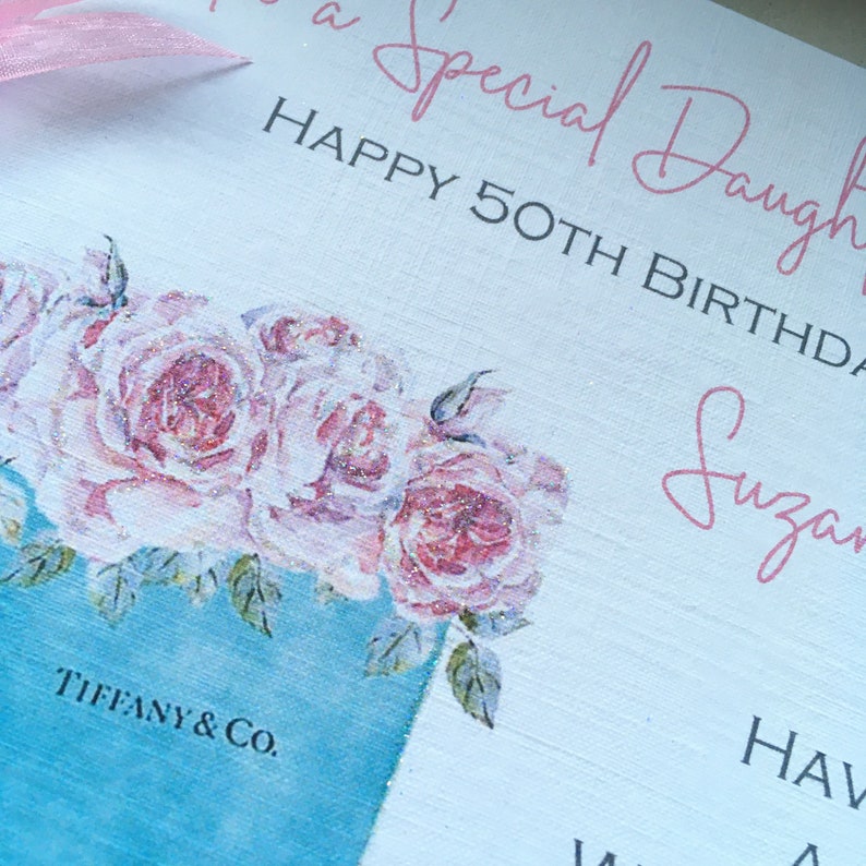Puede incluir: Un letrero decorativo blanco con texto rosa que dice "To a Special Daughter" y "Happy 50th Birthday". El letrero presenta un dise&ntilde;o floral con rosas rosas y una caja de Tiffany & Co.