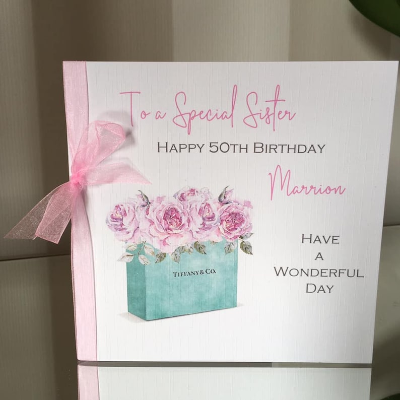 Puede incluir: Una tarjeta de felicitaci&oacute;n blanca con texto rosa y una cinta rosa. La tarjeta dice: "A una hermana especial, Feliz 50 cumplea&ntilde;os, Marrion, Que tengas un d&iacute;a maravilloso". Presenta una ilustraci&oacute;n de acuarela de rosas rosas en una caja turquesa con "Tiffany & Co."