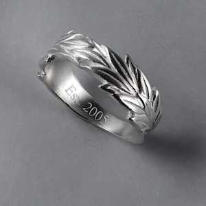 May include: A silver ring featuring a leaf motif. The ring's surface is textured, showcasing intricate leaf patterns. The inscription "Est. 2005" is engraved on the band. The ring is displayed on a grey backdrop.