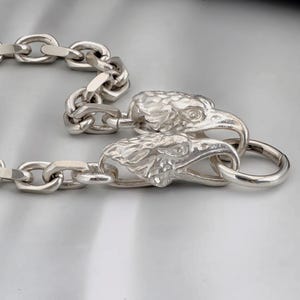 Puede incluir: Un collar de cadena plateada con un cierre de cabeza de águila detallado. Los eslabones de la cadena son de forma ovalada. Las cabezas de águila miran en direcciones opuestas. El collar se muestra sobre una superficie gris claro.