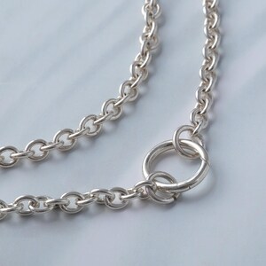 Könnte beinhalten: Eine silberfarbene Halskette mit einem großen kreisförmigen Verschluss. Die Kette besteht aus ovalen Gliedern. Die Halskette wird auf einer hellblauen Oberfläche präsentiert.