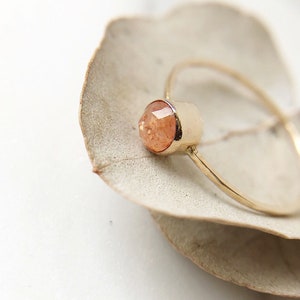 Puede incluir: Un delicado anillo de oro con una pequeña piedra preciosa de piedra de sol facetada. El anillo está colocado sobre un fondo blanco.