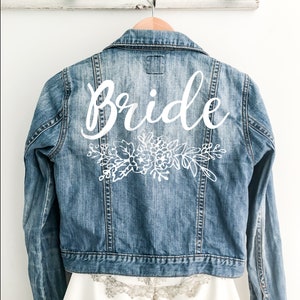 Könnte beinhalten: Eine blaue Jeansjacke mit dem Wort "Bride" in weißer Kursivschrift auf der Rückseite. Das Wort ist mit einem weißen Blumenmuster verziert.