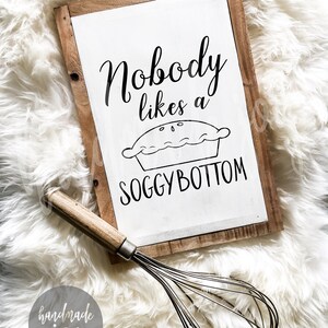 May include: A white wooden sign with black text that reads "Nobody likes a soggy bottom" with a black and white illustration of a pie. The sign is sitting on a white fluffy surface with a silver whisk.