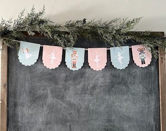May include: A festive banner with alternating pink and blue scalloped flags featuring silhouettes of ballerinas and a nutcracker. The banner is hanging from a garland of evergreen branches.