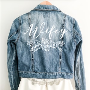 Könnte beinhalten: Eine blaue Jeansjacke mit dem Wort "wifey" in weißer Schrift auf der Rückseite. Unter dem Wort befindet sich ein weißes Blumenmuster. Die Jacke hängt an einem Holzbügel.