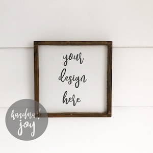 May include: A white wooden sign with a brown wood frame. The sign says "your design here" in gray cursive lettering. The sign is hanging on a white wall.