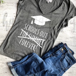 May include: Gray t-shirt with a white graduation cap and the text "Schools Out For Summer Forever!" printed on it. The shirt is laying on a wooden surface with a pair of blue jeans.