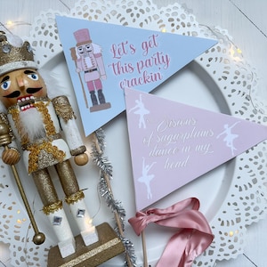 Puede incluir: Una escena festiva con un cascanueces de madera dorada y dos banderines triangulares. Uno es azul claro con una ilustración de cascanueces y el texto "Let's get this party crackin'". El otro es rosa con siluetas de bailarinas y el texto "Visions of sugarplums dance in my head."